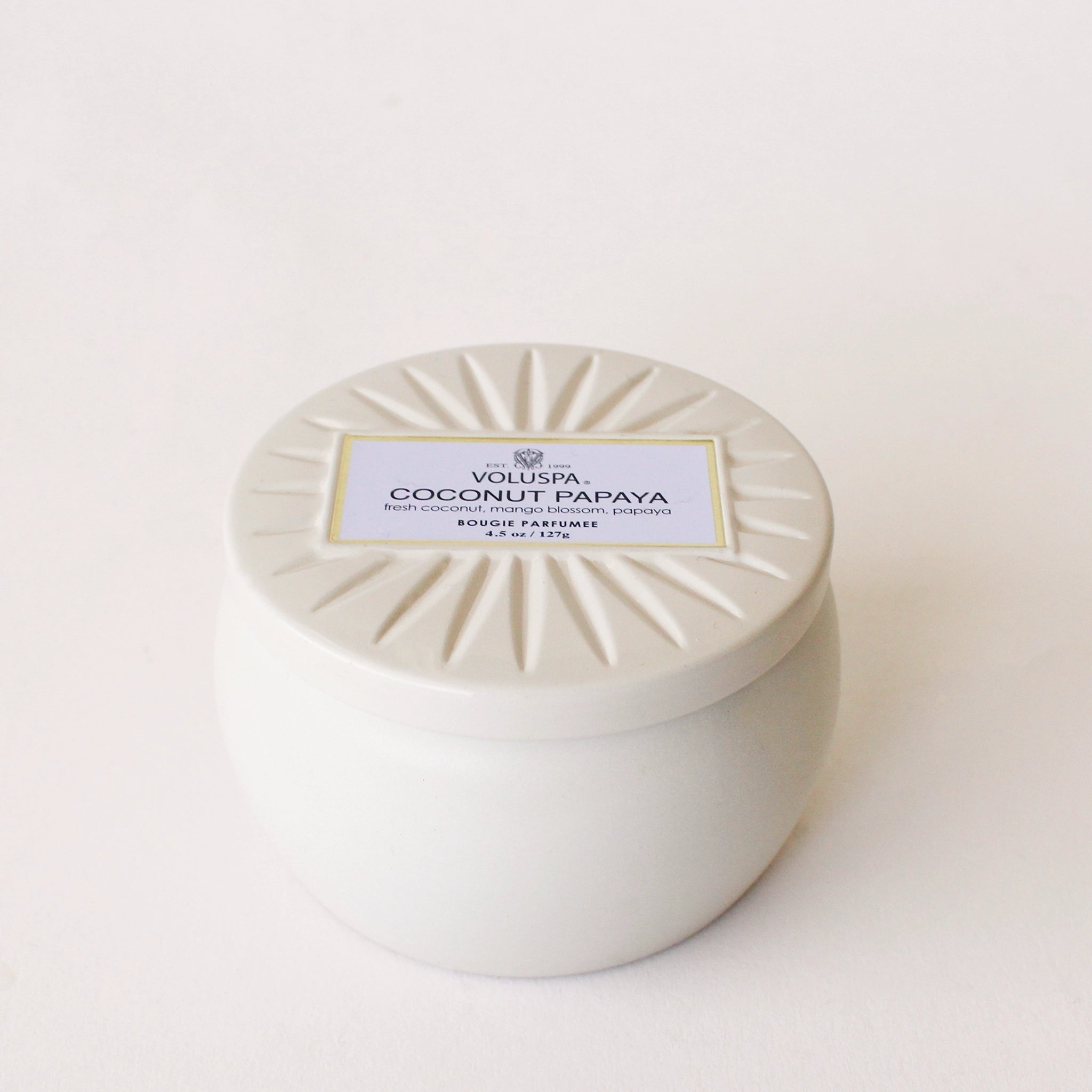 On a cream background is a round tin candle with a lid and a single wick candle inside and a label that reads, "Voluspa Coconut Papaya".