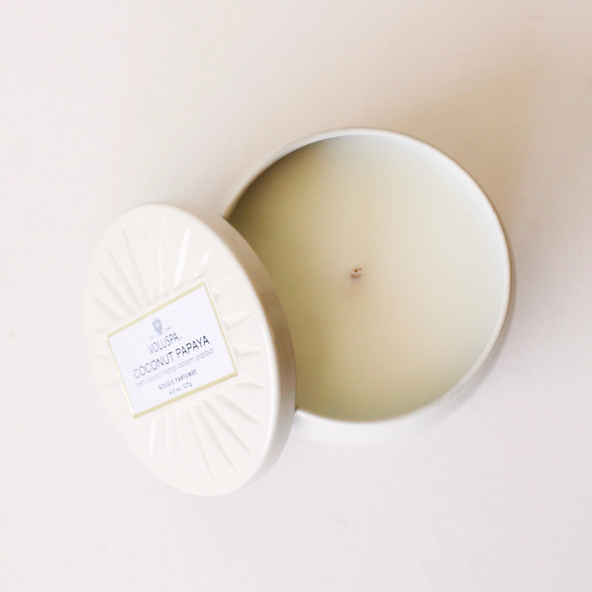 On a cream background is a round tin candle with a lid and a single wick candle inside.