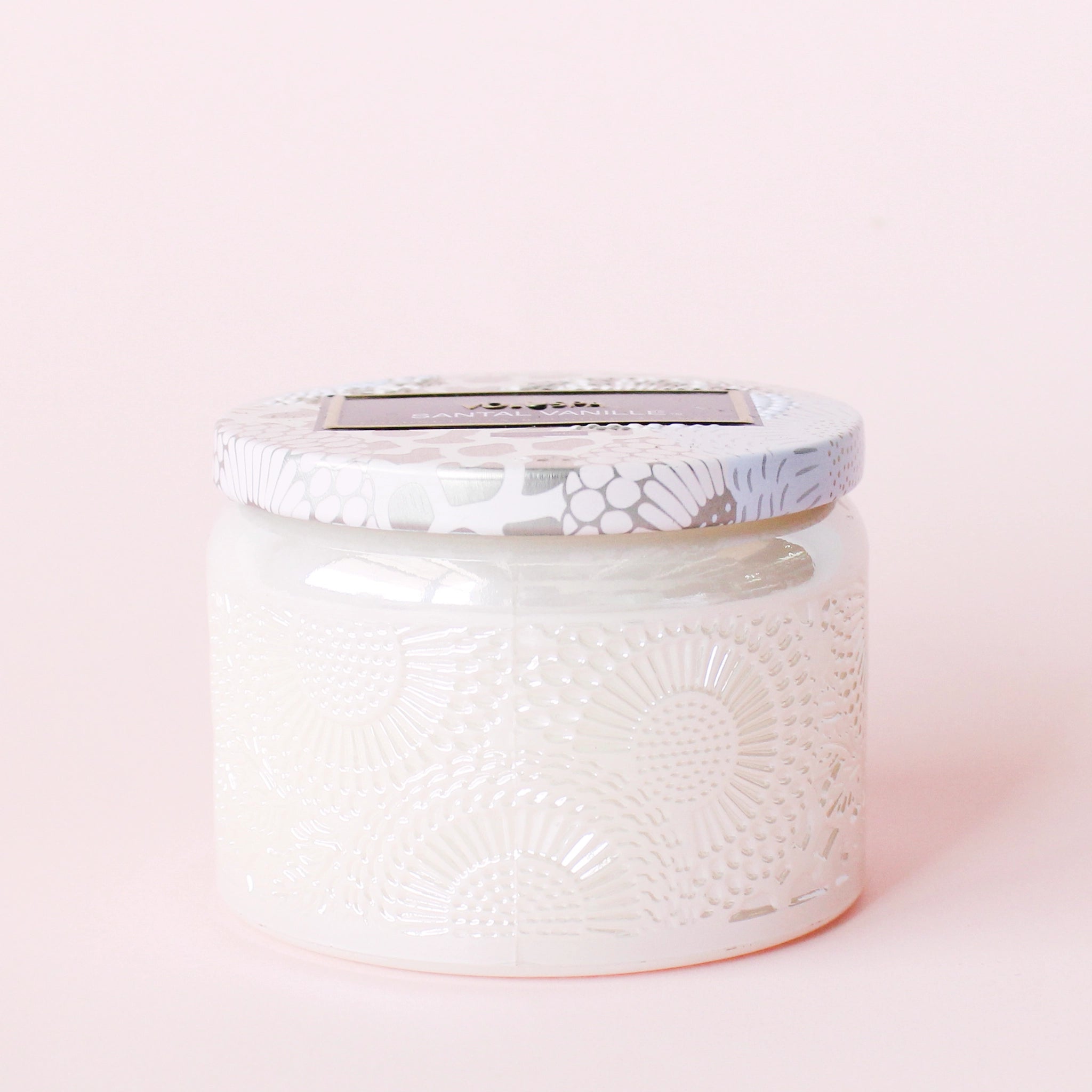 In front of a pink background is a light pink glass jar. The jar has a floral pattern on it. On top of the candle is a tin lid. The lid is light pink, lavender and silver floral pattern. In the middle is a black rectangular sticker.