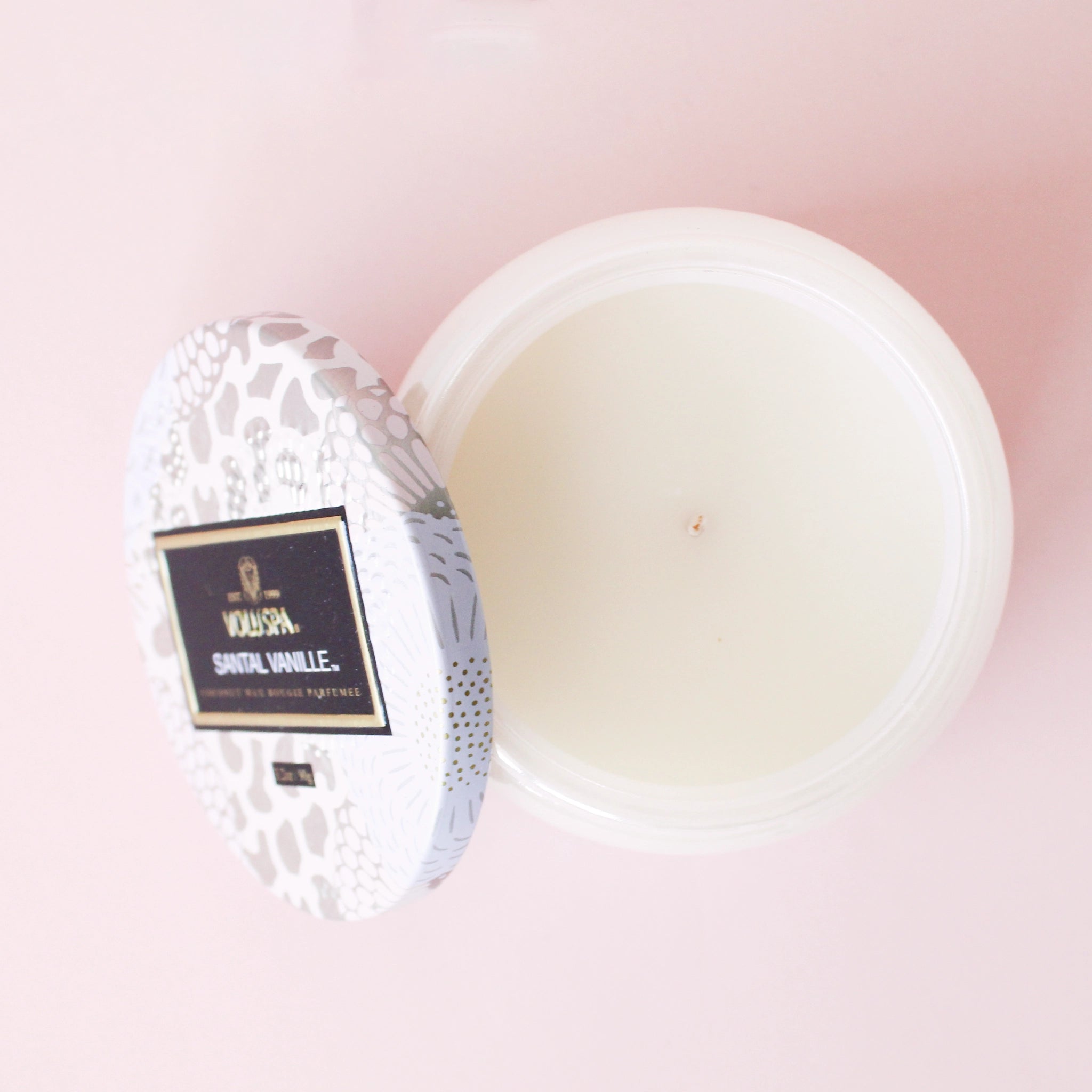 Birds eye view of a cylinder candle. The candle is white with a white wick in the center. Leaning against the left side is the round tin lid. The lid is light pink, lavender and silver floral pattern. In the middle is a black rectangular sticker. Inside the sticker is gold text that reads ‘voluspa.’ Below is white text that reads ’santal vanille.’