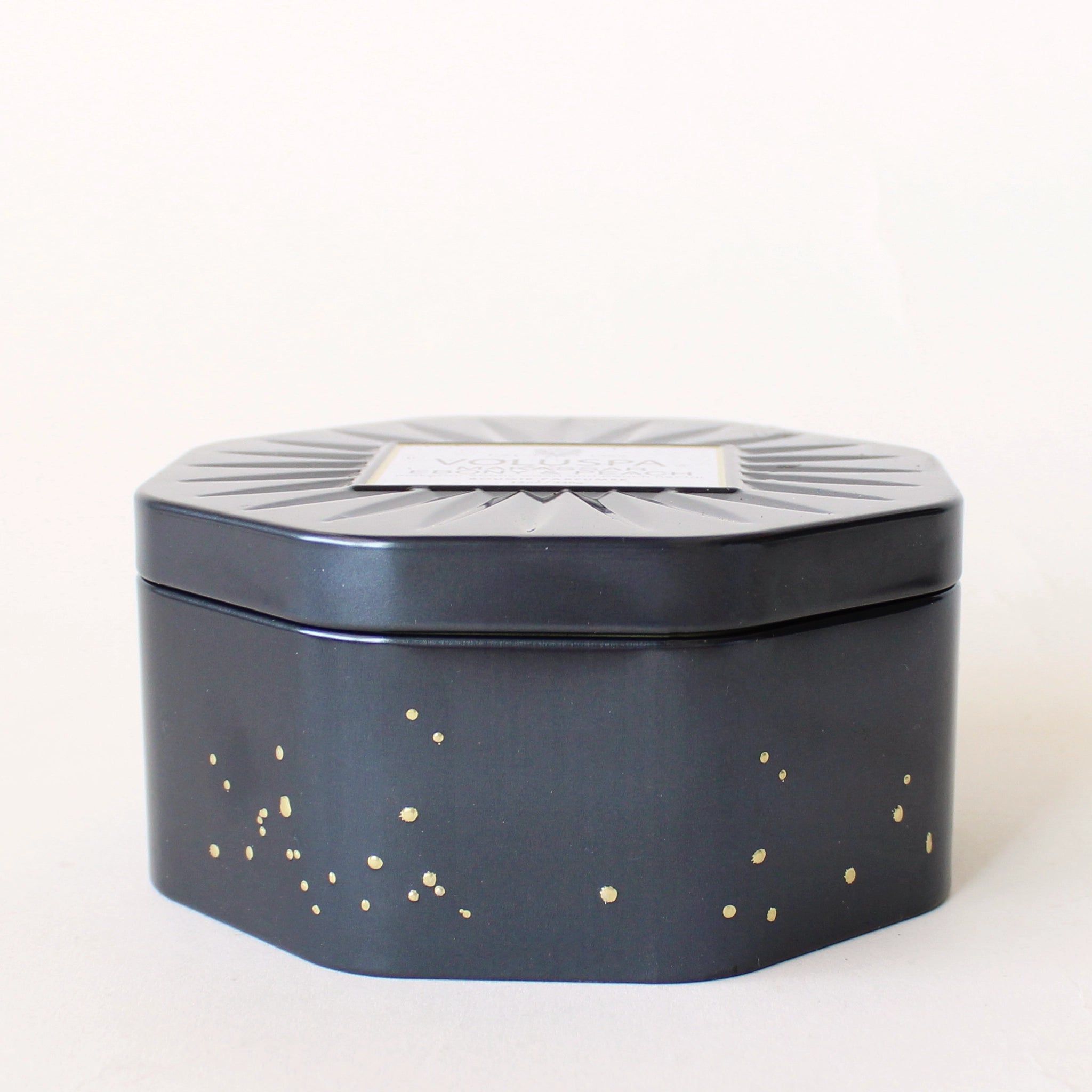 On a white background is an octagon shaped tin candle with three wicks, white wax and small gold speckle detailing.