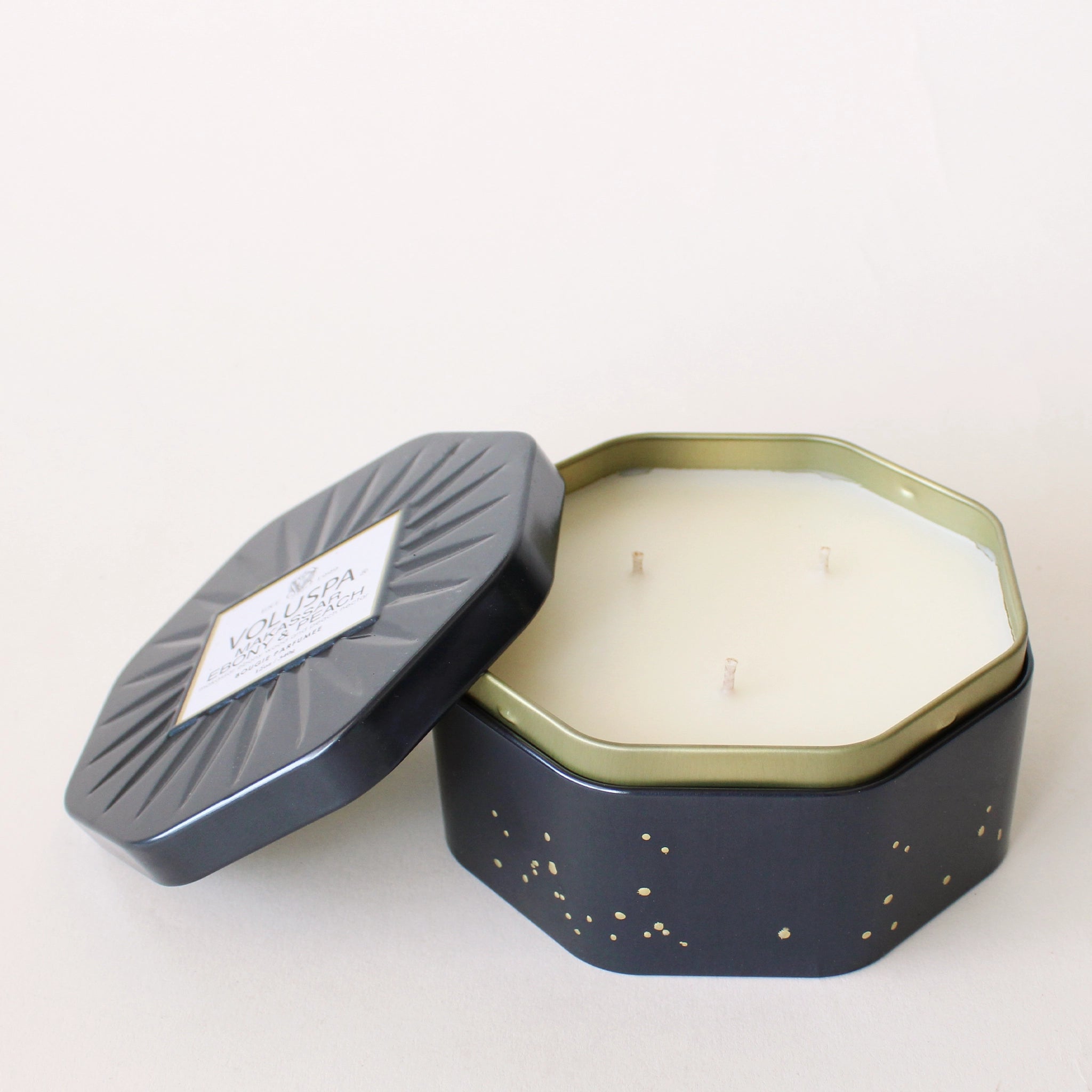 On a white background is an octagon shaped tin candle with three wicks, white wax and small gold speckle detailing. 