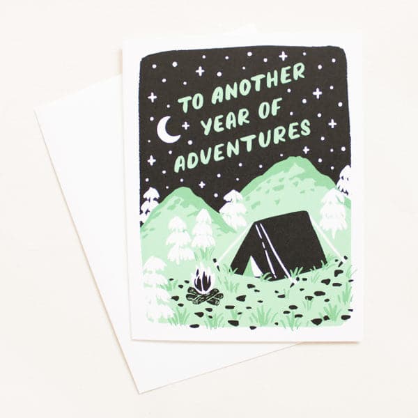An illustrated greeting card with white envelope. The illustration is a night scene of a black tent nestled in a forest of white trees and green hills. Mint green text says &quot;To Another year Of Adventures.&quot;