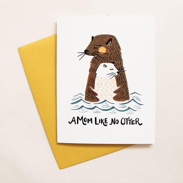 A white card with the text &quot;a mom like no otter&quot; along with yellow envelope. Illustration shows a brown otter hugging a baby white otter while in water. 