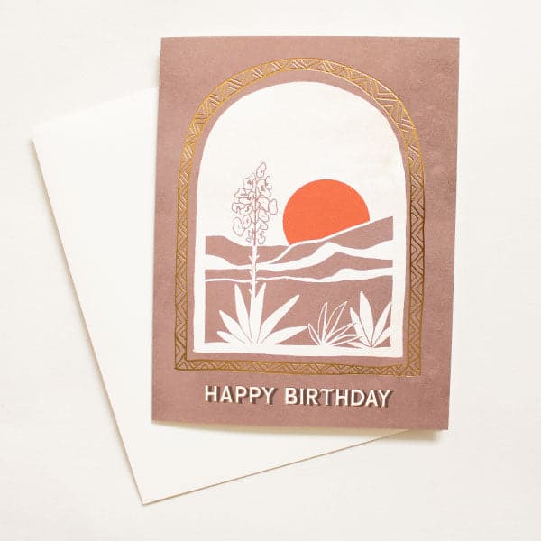 A brown card with a desert illustration and aloe plants along with an orange sun and an arch design with white text on the bottom that reads, "Happy Birthday". Also included is a white envelope.