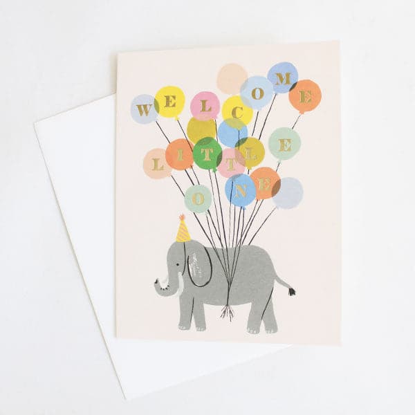 This playful cream colored card features a sweet great elephant wearing a festive yellow and pink party hat. Th elephant is tied to a bundle of colorful birthday balloons reading 'Welcome Little One'. Each golden letter is homed in a separate balloon. The card is accompanied by a solid white envelope.  
