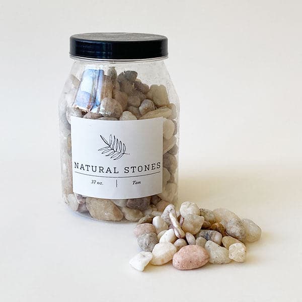 On a cream background is an assortment of natural tan and cream colored stones in a plastic jar with a black lid.
