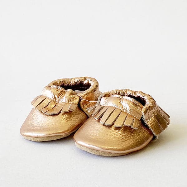 In front of a white background is a pair of gold baby moccasins. The shoes have a garland like border on the front top and the back of the shoe.