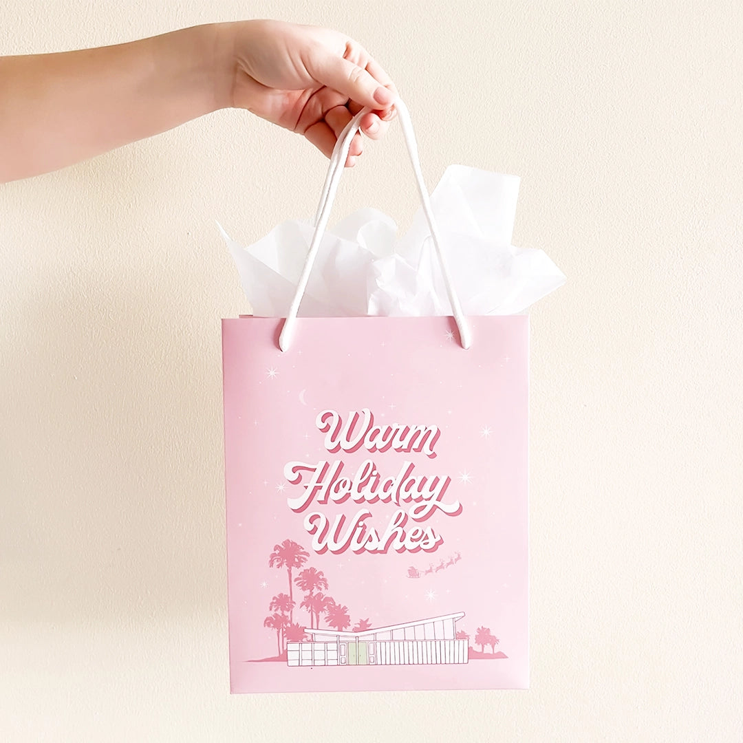 A cool toned pink paper gift bag with a graphic of a midcentury home in front of palm trees along with text that reads, "Warm Holiday Wishes" on the large size and "Merry Christmas" on the smaller one.