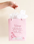 A cool toned pink paper gift bag with a graphic of a midcentury home in front of palm trees along with text that reads, "Warm Holiday Wishes" on the large size and "Merry Christmas" on the smaller one.