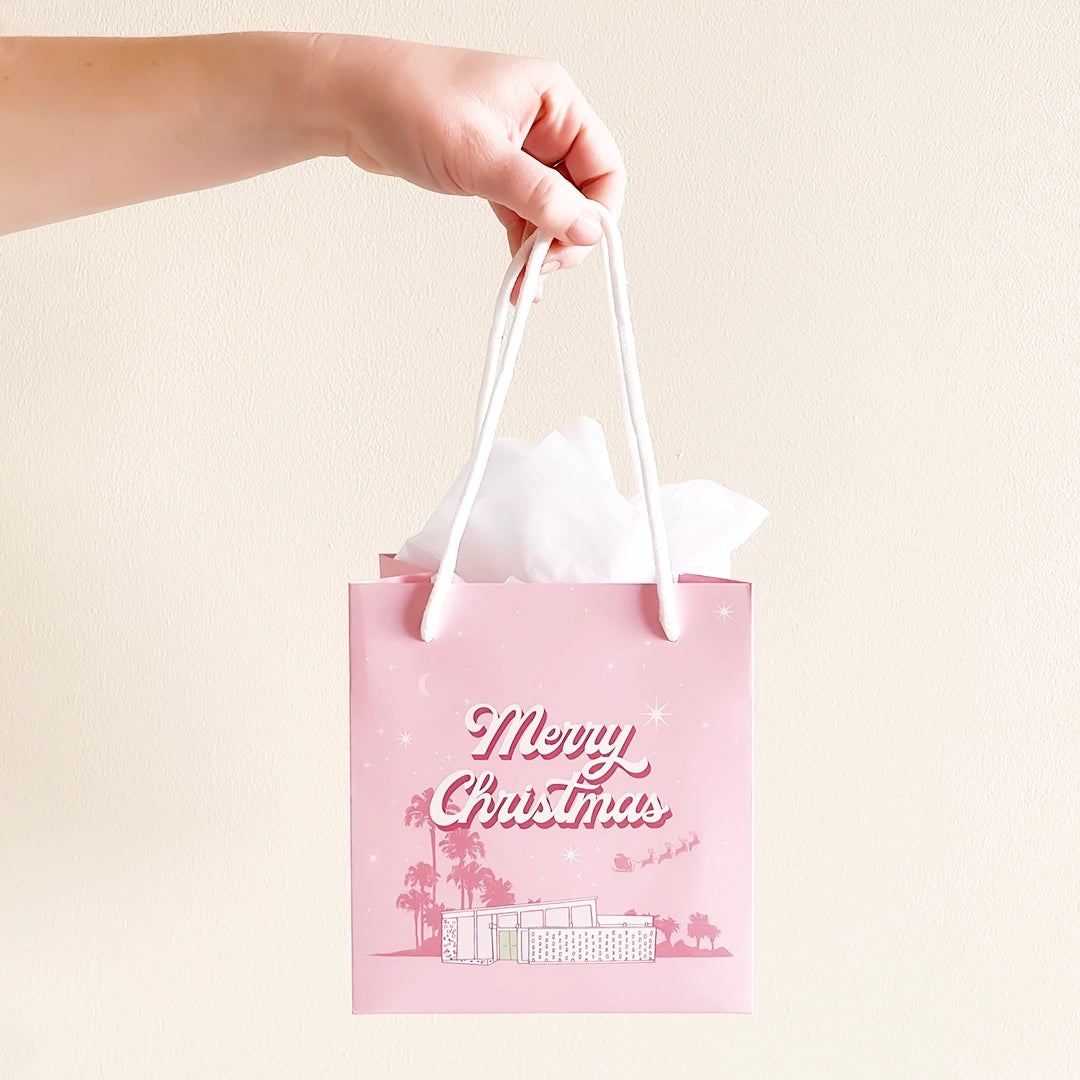 A cool toned pink paper gift bag with a graphic of a midcentury home in front of palm trees along with text that reads, "Warm Holiday Wishes" on the large size and "Merry Christmas" on the smaller one.