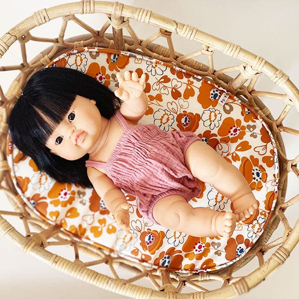 An asian baby doll with black straight hair and dressed in a pink onesie and sitting inside of a rattan crib. 