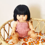 An asian baby doll with black straight hair and dressed in a pink onesie and sitting inside of a rattan crib. 