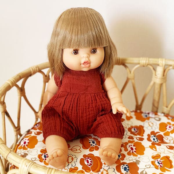 Baby doll toy with a blonde bob haircut in a deep red romper outfit that is sold separately. Doll is positioned on bohemian rattan crib, sitting upright. 