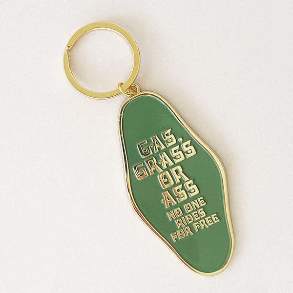 On a white background is a diamond shaped metal keychain that reads, "Gas, Grass or Ass No Ones Rides For Free" along with a gold loop at the top to attach to keys. 