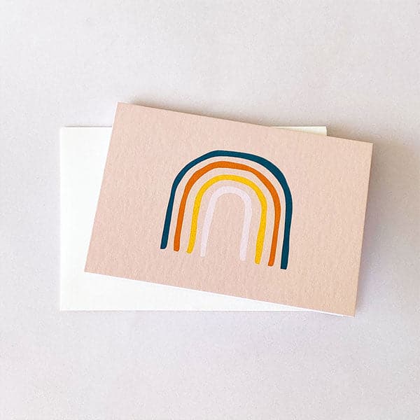 On top of a white envelope is a horizontal pink card. In the middle is an arched rainbow. The colors of the rainbow are blue, orange, yellow and light pink. 
