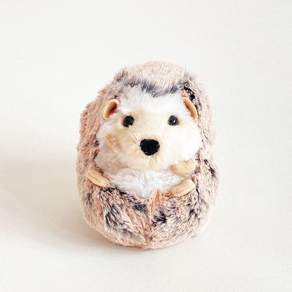 In front of a white background is a fluffy ball stuffed animal. It has black and brown marbled fur with a white chest and face. It has four tan little feet on its chest. There are two black eyes and one black nose. At the top there are two tan ears.