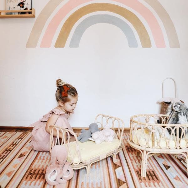 This is a picture of a little girl wearing a light pink dress. She is kneeling down, playing with her toys. In front of her is a light tan wicker day bed. It arches on the right and left side of the day bed. Inside the day bed is a yellow mattress. Inside the day bed is two stuffed animals. 