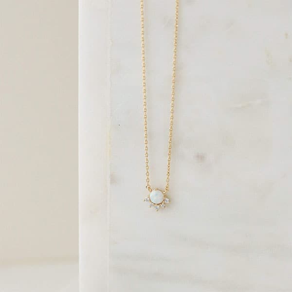 A dainty gold necklace with a round opal pendant in the center that is lined with cz stones around the bottom edge. 