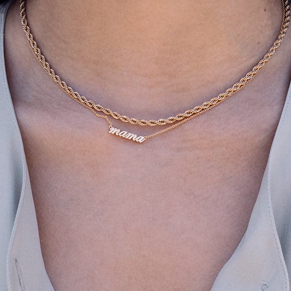 Zoomed in photo of a woman&#39;s neck. She is wearing two gold necklaces. One is a thick rolled gold chain and the other necklace reads &quot;mama&quot; in lowercase script. A thin chain is connected on each side of the word. 