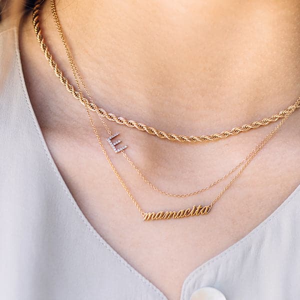 Zoomed in photo of a woman's neck. She is wearing 3 gold necklaces layered on her neck. The top one is a thick rolled gold chain, underneath that is a thin gold chain with a silver and diamond capitol letter "E" and the other necklace reads "mamacita" in lowercase script. 
