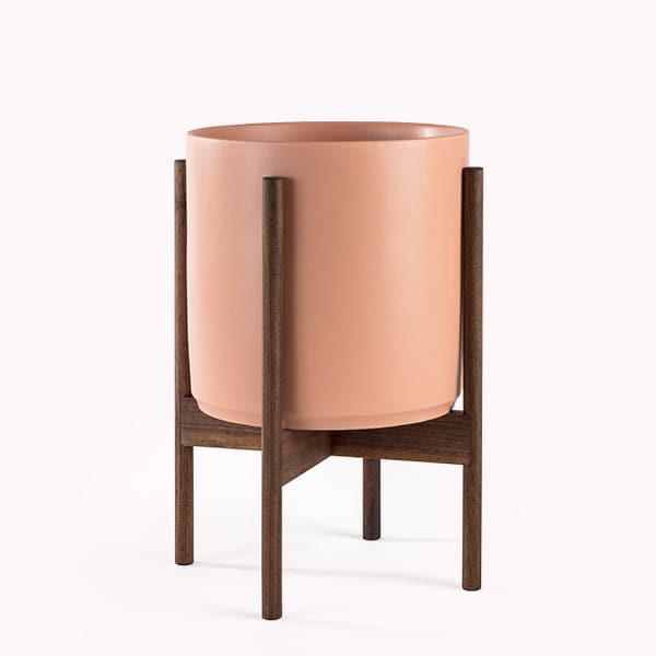 This cylinder pot is a peachy color and sits within four spokes of a walnut wood plant stand, standing about 7 inches from the ground.