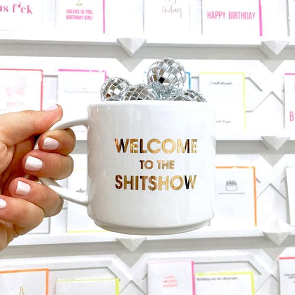 Classic white ceramic mug with a thin round handle labeled 'Welcome to the Shitshow' in reflective gold lettering. Mug is held by a freshly manicured hand and is filled with shimmering miniature disco ball. Background is a card stand displaying white cards labeled 'Happy Birthday'