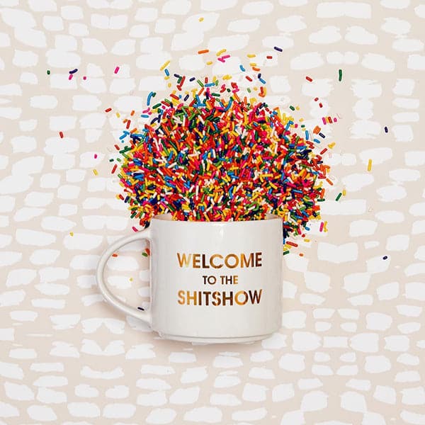 Classic white ceramic mug with a thin round handle labeled 'Welcome to the Shitshow' in reflective gold lettering. Mug is positioned pouring colorful rainbow sprinkles across a boho white and cream backdrop.