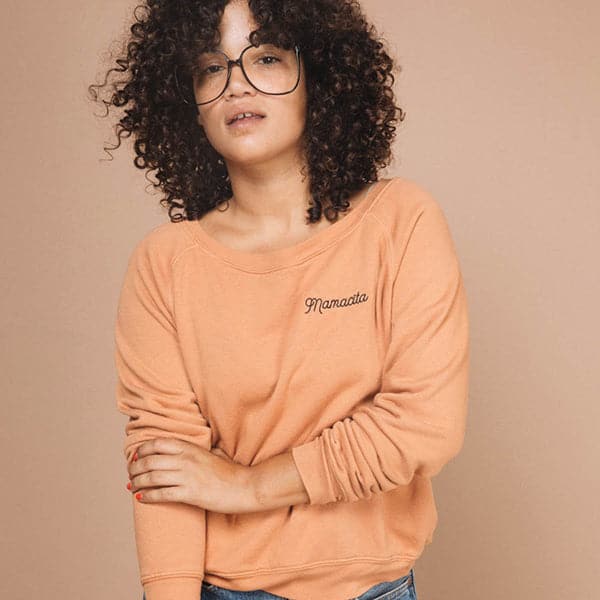 A crew neck sweatshirt in a light rust color with black embroidered letters in the right corner that reads, "Mamacita" worn here on a model in front of a peachy background. 
