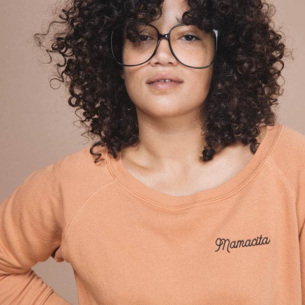 A crew neck sweatshirt in a light rust color with black embroidered letters in the right corner that reads, "Mamacita" worn here on a model in front of a peachy background.