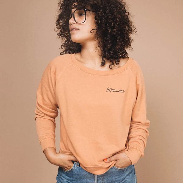 A crew neck sweatshirt in a light rust color with black embroidered letters in the right corner that reads, "Mamacita" worn here on a model in front of a peachy background.