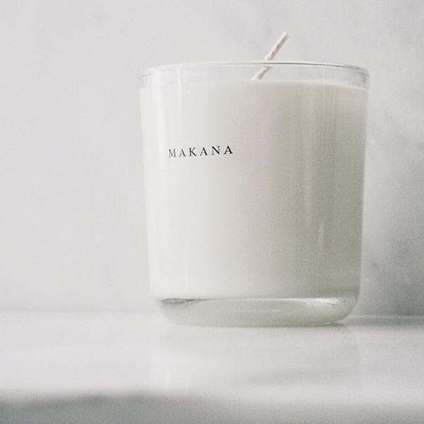 In front of a white and gray marbled wall is a round glass jar. Inside the jar is a white candle with a white wick in the center. On the front is black text that reads ‘Makana.’ 