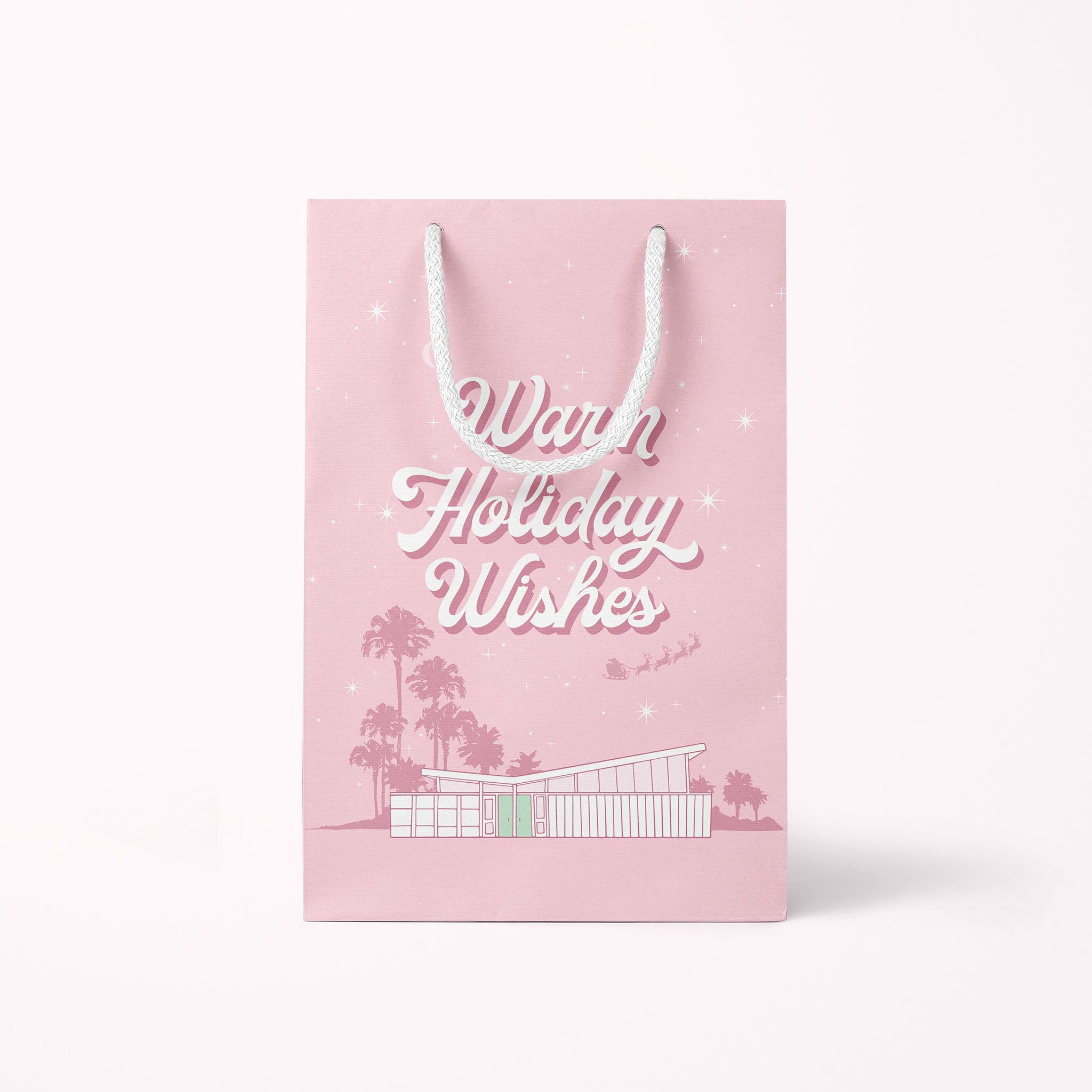 A cool toned pink paper gift bag with a graphic of a midcentury home in front of palm trees along with text that reads, "Warm Holiday Wishes" on the large size and "Merry Christmas" on the smaller one.