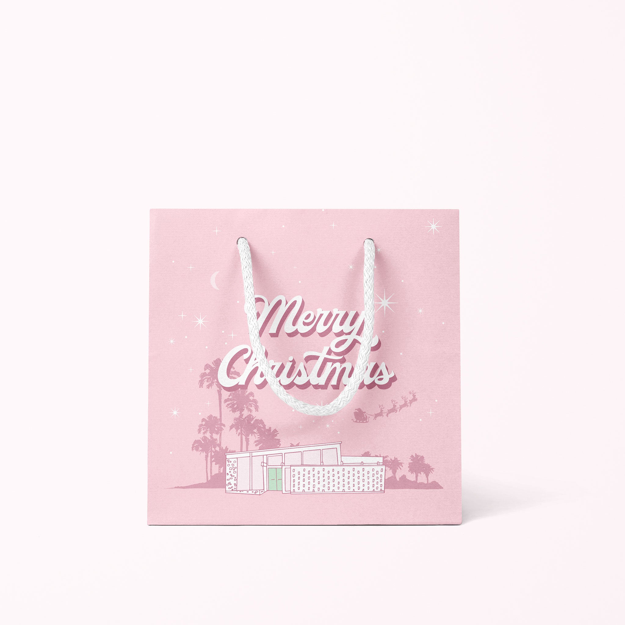 A cool toned pink paper gift bag with a graphic of a midcentury home in front of palm trees along with text that reads, "Warm Holiday Wishes" on the large size and "Merry Christmas" on the smaller one.