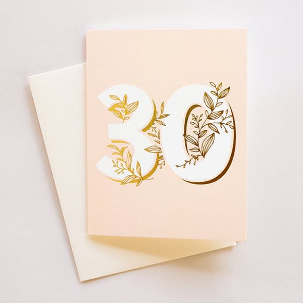 A light pink card with a cream colored envelope and big white numbers that read, "30" along with gold foiled dainty vines that wrap through and around the numbers.