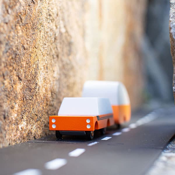 On the left style of the picture is a dark brown rock wall. On the ground is a black rode with a white dividing line. Next to the rock wall is a red wood card with a white top and two black lines down the side. Behind it is an orange and white trailer. 