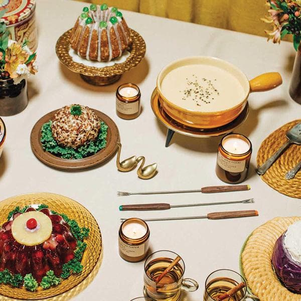 Birds eye view of table with a white table cloth. Spread out across the table is plates and dishes with different types of festive foods. There's a red gelatin mold and a white ball of cheese covered with brown nuts. Above the cheese is a gold dish with a brown bundt cake with white icing. To the right is a gold dish filled with melted cheese. There are three dark amber, cylinder glass jars spread out between the dishes. They are all filled with a white candle and the wick is lit. 