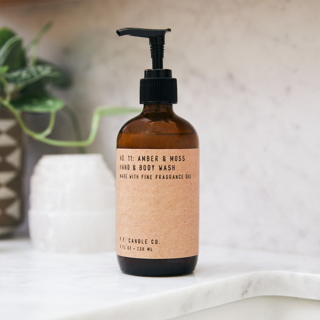Translucent brown hand soap dispenser on a marble bathroom candle, with brown paper label sticker "No. 11 Amber and Moss, Hand and Body Wash. Made with Fine Fragrance Oil. P.F. Candle Co."