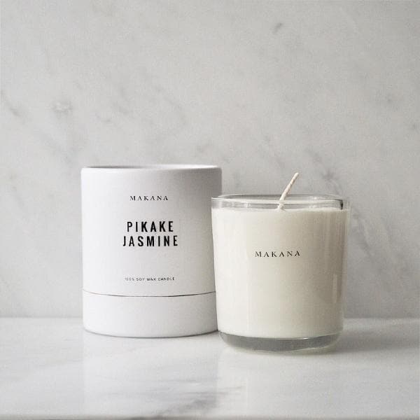 In front of a white and gray marbled wall is a round glass jar. Inside the jar is a white candle with a white wick in the center. On the front is black text that reads ‘Makana.’ To the left is a white round package. There is black text on the front that reads ‘Makana Pikake Jasmine.’ 