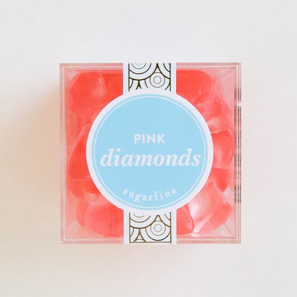 In front of a white background is a clear cube container. The container is filled with light pink diamond shaped gummies. In the middle is a bright blue round sticker with a white border. In the middle is white text that reads ‘pink diamonds.’ 