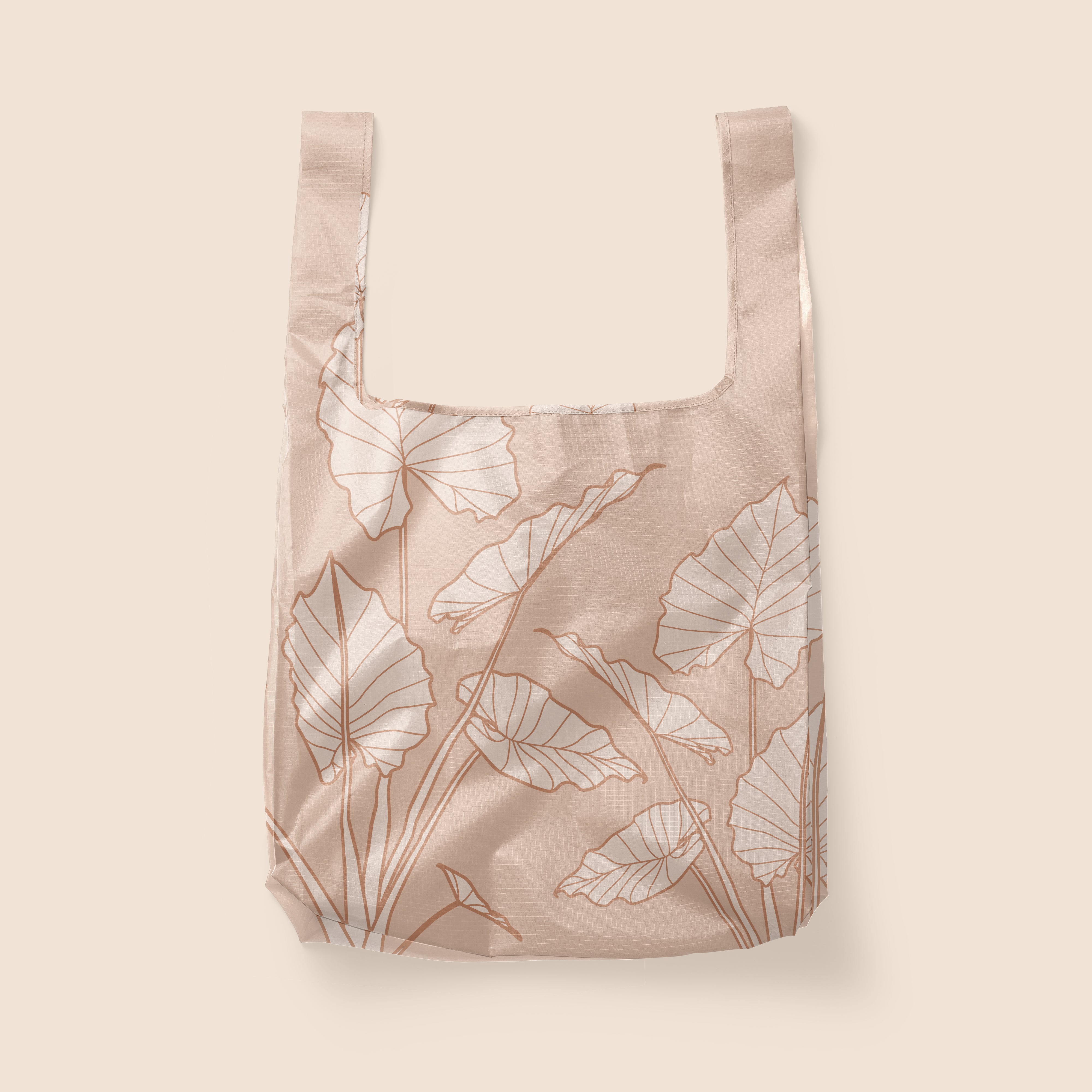 Photographed in front of a white background is a light pink / tan nylon reusable tote with cream and darker terracotta line drawings of alocasia leaves. There is a squared off design to the bag and two handles that can be held or used over your shoulder.