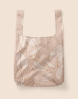 Photographed in front of a white background is a light pink / tan nylon reusable tote with cream and darker terracotta line drawings of alocasia leaves. There is a squared off design to the bag and two handles that can be held or used over your shoulder.