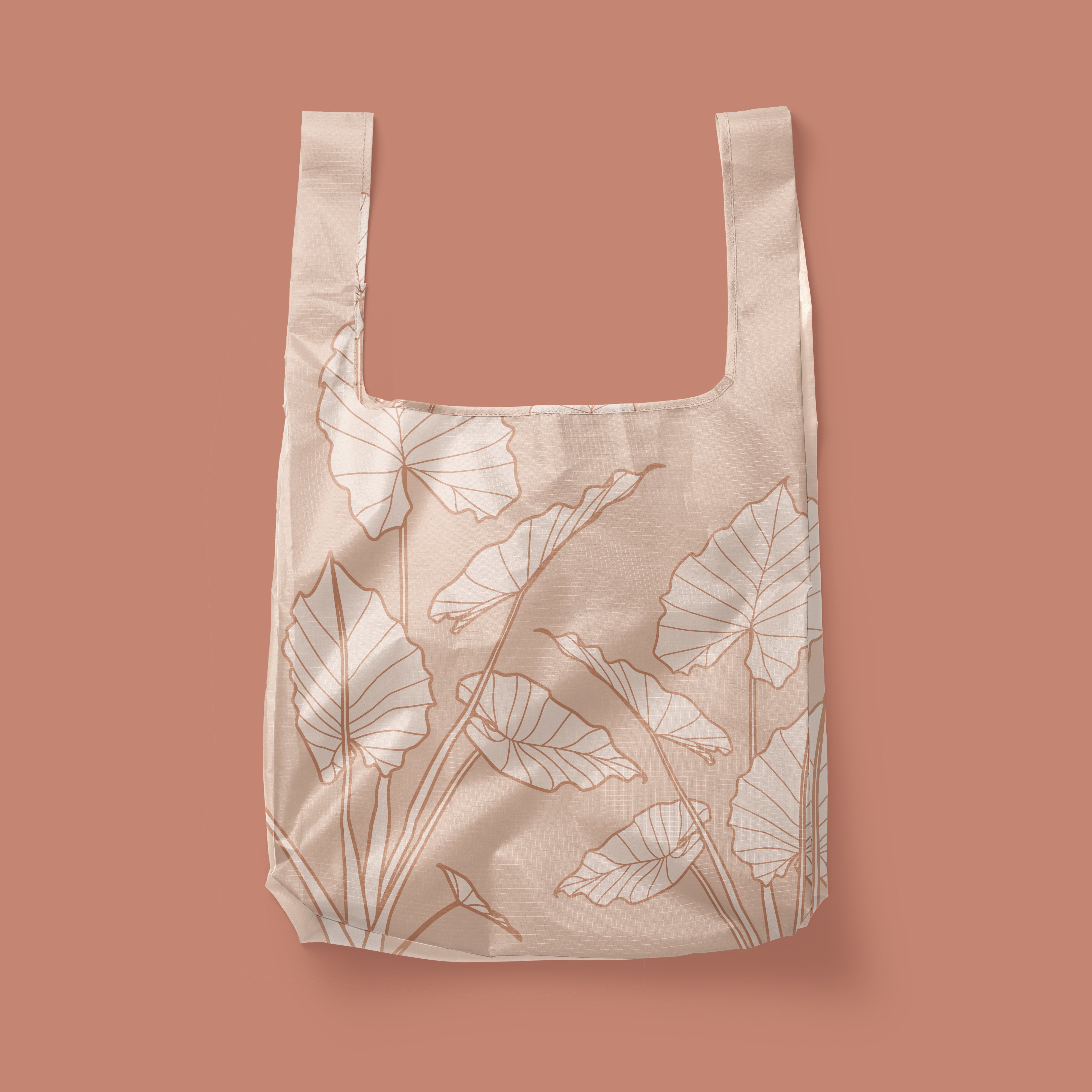 Photographed in front of a rose colored background is a light pink / tan nylon reusable tote with cream and darker terracotta line drawings of alocasia leaves. There is a squared off design to the bag and two handles that can be held or used over your shoulder.