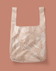 Photographed in front of a rose colored background is a light pink / tan nylon reusable tote with cream and darker terracotta line drawings of alocasia leaves. There is a squared off design to the bag and two handles that can be held or used over your shoulder.