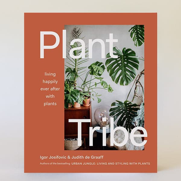 In front of a white background is a book cover. The book is a rust colored. On the right side is a picture of a home with dark green plants on a wood table. Across the top and bottom of the book is white text that reads ‘plant tribe.’ To the left of the picture is white text that reads ‘living happily ever after with plants.&#39;