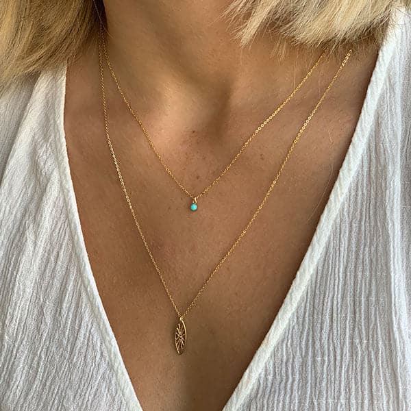 This is a picture of a woman’s neck and chest. She has short blonde hair and is wearing a white v-neck blouse. Around her neck is a thin gold chain necklace with a small turquoise bead in the middle. She is wearing a second thin gold chain necklace that falls below her collar bone. There is an oval shaped, gold pendent in the middle.