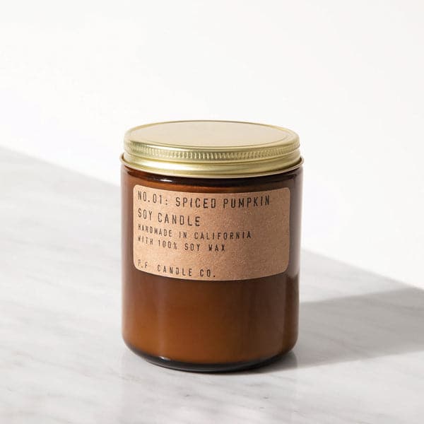 In front of a white background is a dark amber, cylinder glass jar. On top is a gold, metal lid. On the top, front of the candle is a light brown sticker. On the sticker is black text that reads ‘No 01 spiced pumpkin soy candle made in California with 100% soy wax. PF candle co.'