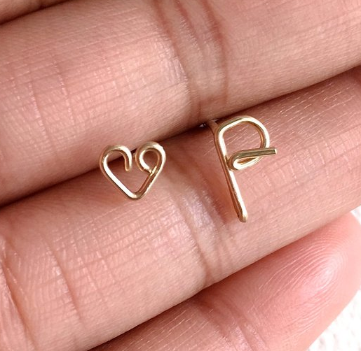 A hand holding two stud earrings of bent gold wire. One in the shape of a heart and one in the shape of the letter P.