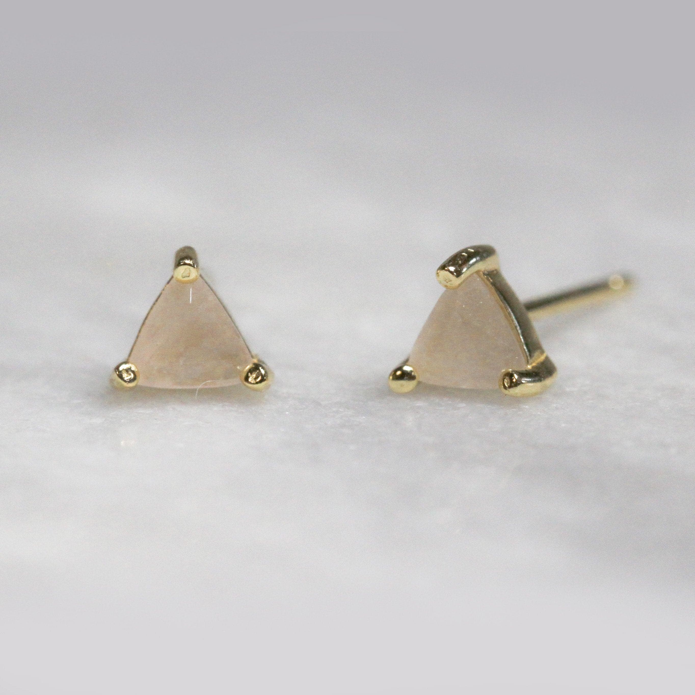 In front of a white background is a pair of rose quartz studs. The crystal is shaped like a triangle with a gold post on each corner. 