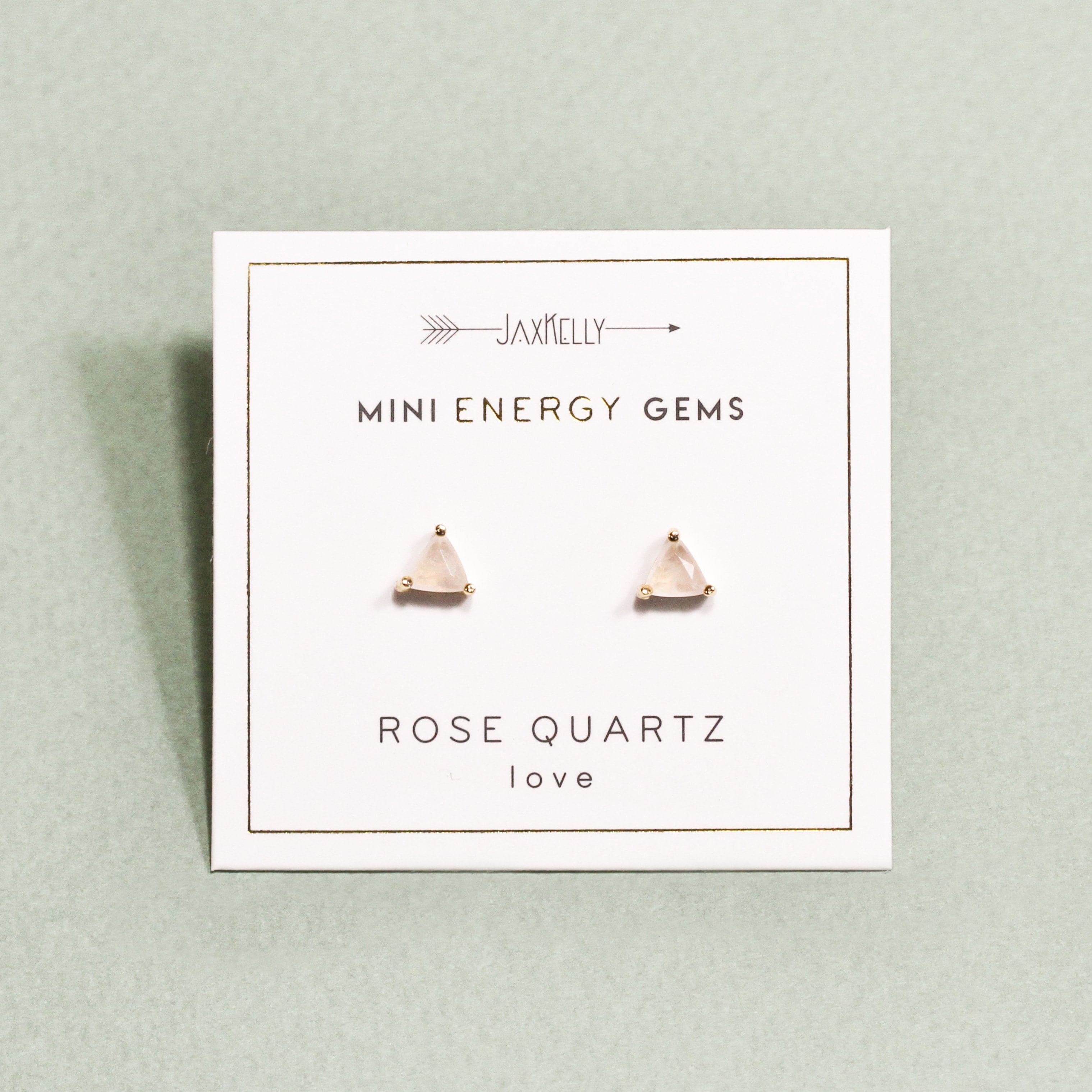 In front of a gray background is a white square card. About an inch in is a gold border. In the middle is a pair rose quartz studs. The studs are shaped like a triangle with a gold post on each corner. Above is gold text that reads ‘mini energy gems.’ Below is more gold text that reads ‘rose quartz love.’ At the top of the car is a gold arrow. In the middle of the arrow is gold text that reads ‘Jaxkelly.&#39;